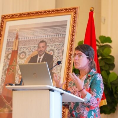 Woman giving a speech, standing in front of a flag and a portrait.
