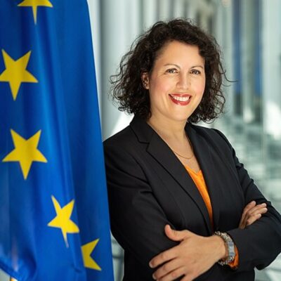 Woman standing besides the European Union flag and looking in the camera.